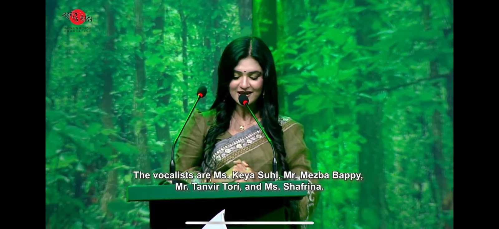 Sanjida Rahman speaking at a podium with a forest-themed screen behind her